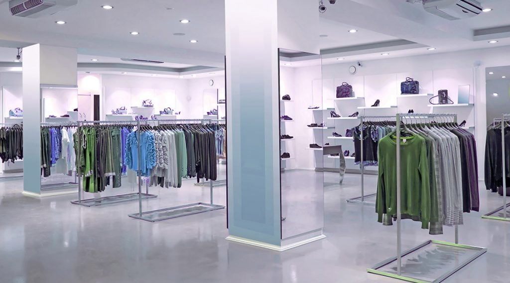 A modern, brightly lit clothing store featuring racks of neatly arranged clothes in green and blue, with shoes, handbags, and accessories displayed against white walls and sleek epoxy flooring popular in Palm Beach and Miami-Dade.
