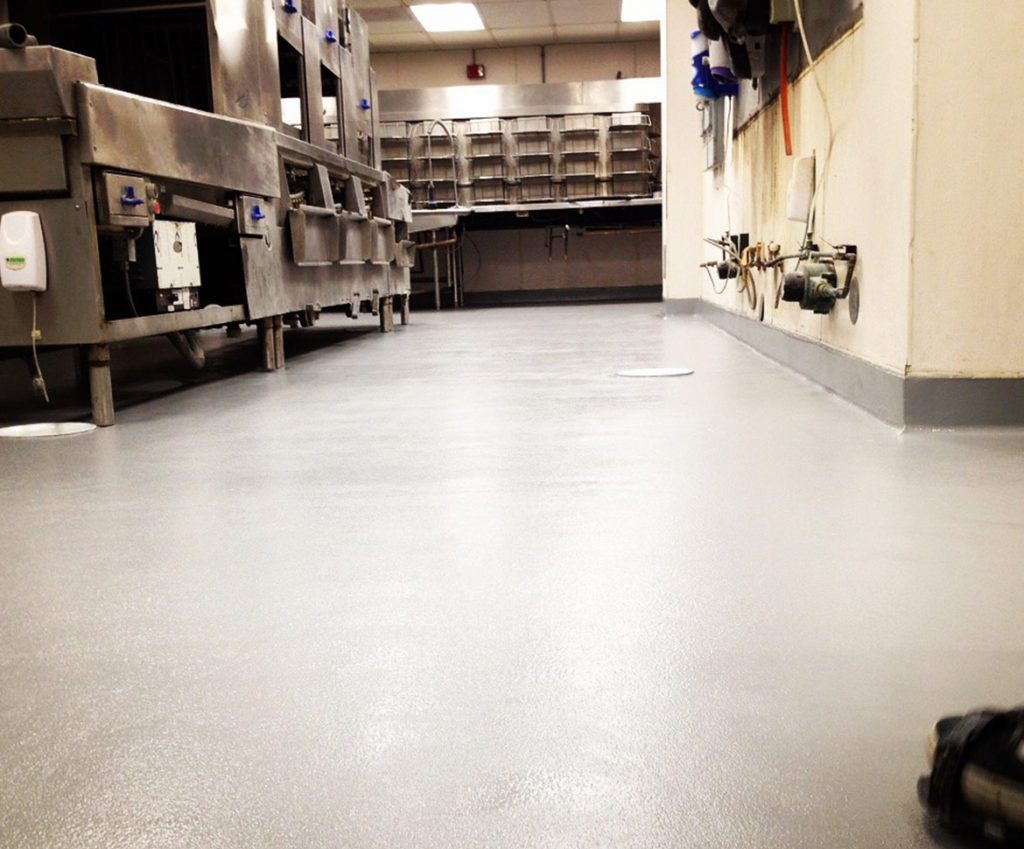 Commercial kitchen with a shiny, clean, gray epoxy flooring Palm Beach finish, stainless steel equipment on the left, sinks and shelves at the back, and various pipes and utilities lining the right wall.