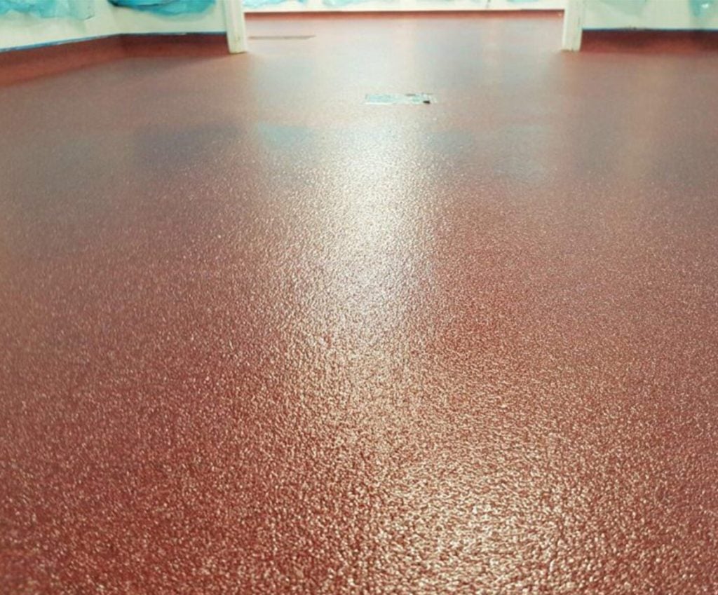 Close-up view of a textured reddish-brown epoxy flooring Palm Beach surface, with slightly blurred white walls and blue coverings visible in the background.