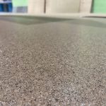 Close-up view of a speckled, textured surface in shades of brown, beige, and black—likely epoxy flooring Palm Beach style—with a blurred background showing parts of a room and green and beige walls.