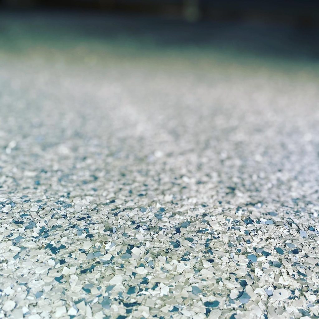 A close-up view of a textured epoxy flooring Palm Beach surface, covered with small, light-colored flakes that create a rough, sparkling appearance. The blurry background draws focus to the intricate detail of this popular Broward, FL flooring style.