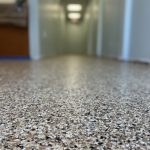 Low-angle view of a speckled, textured epoxy flooring in a hallway with pale green walls and ceiling lights, leading toward a blurred background with indistinct office furniture—typical of spaces in Palm Beach and St. Lucie County, FL.