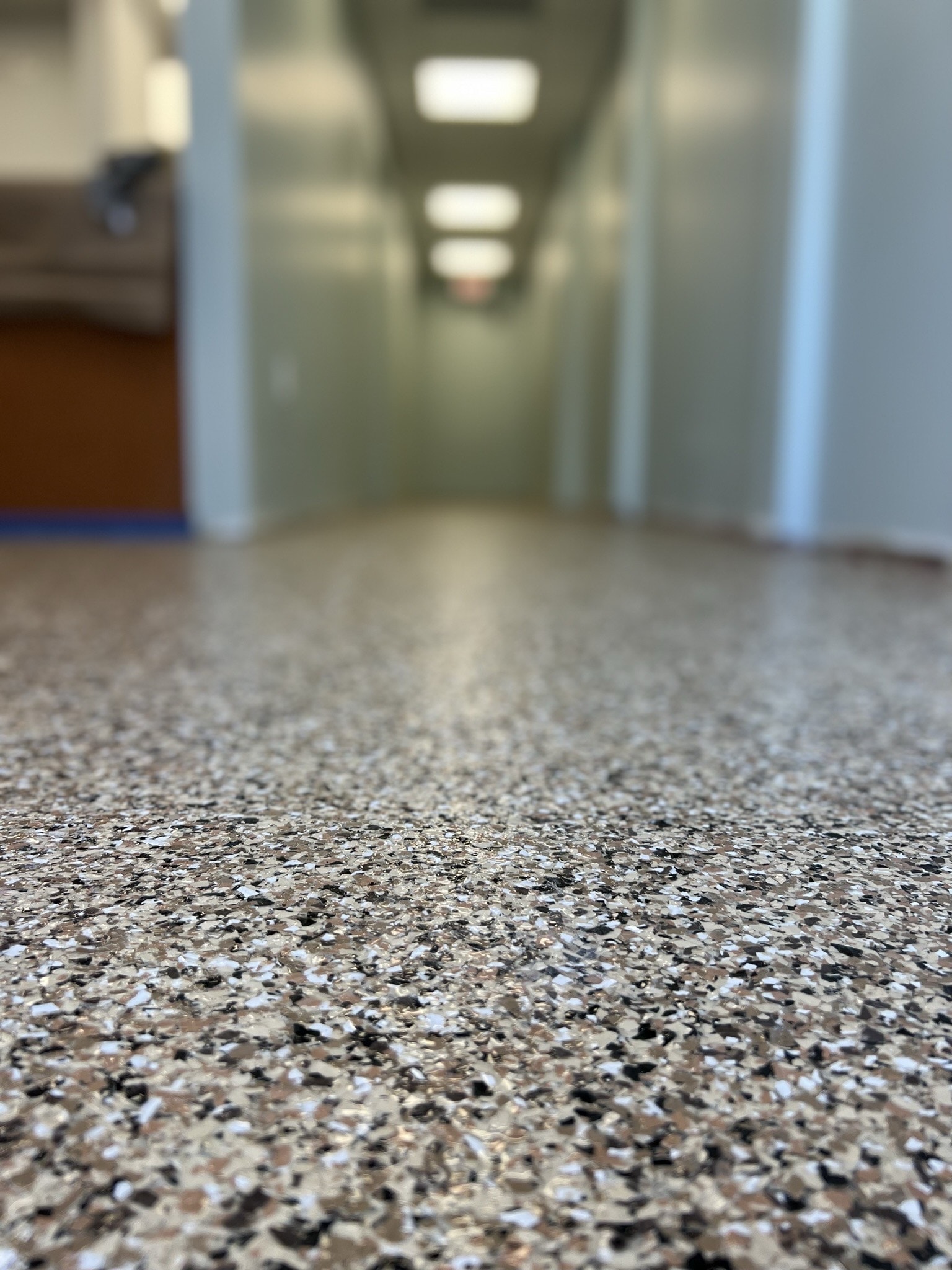 Low-angle view of a speckled, textured epoxy flooring in a hallway with pale green walls and ceiling lights, leading toward a blurred background with indistinct office furniture—typical of spaces in Palm Beach and St. Lucie County, FL.