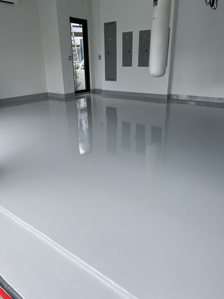 A clean, empty room with smooth, shiny gray epoxy flooring Palm Beach and St. Lucie County, FL; white walls, several electrical panels, a door, and a hanging punching bag visible in the corner.