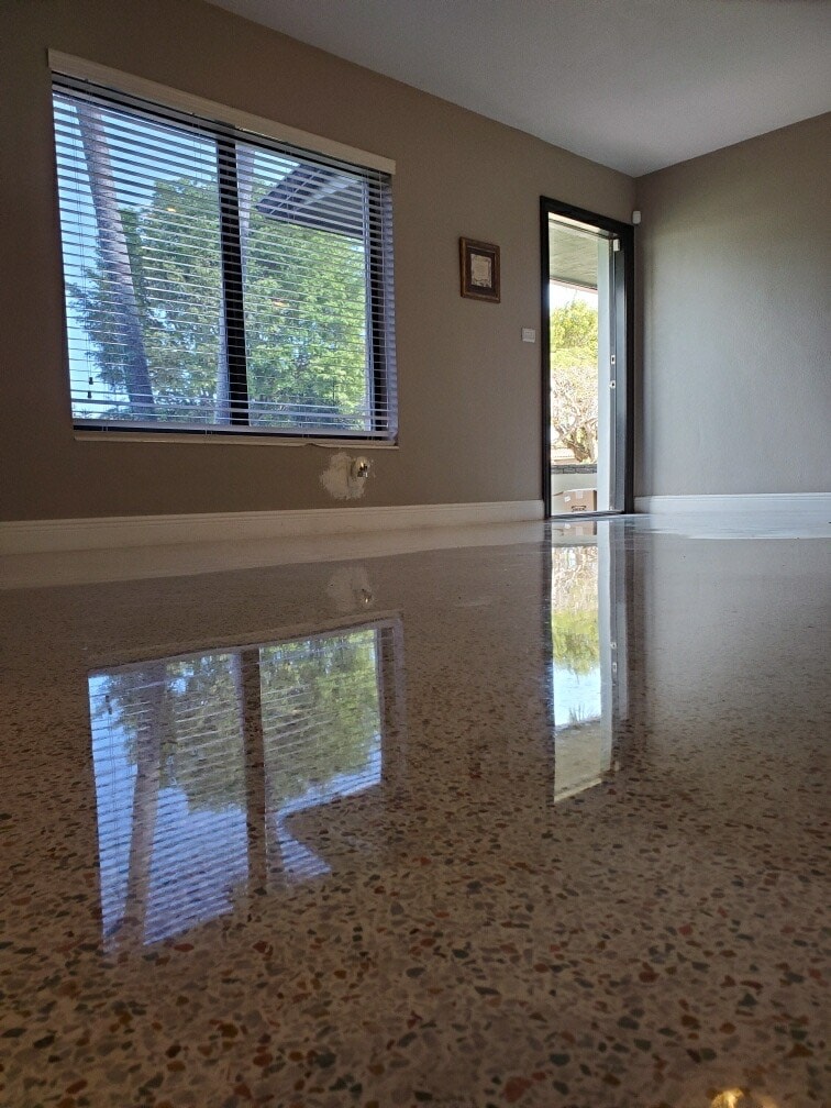 A highly polished terrazzo floor reflects a window with blinds and an open door in a mostly empty room with beige walls and white trim, letting in natural light—an elegant look popular in Miami-Dade, FL, and St. Lucie County homes.