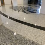 A polished epoxy flooring Palm Beach, FL features a large black geometric star center design, surrounded by curved black lines. Light terrazzo covers most of the area, with darker speckled flooring in the foreground.