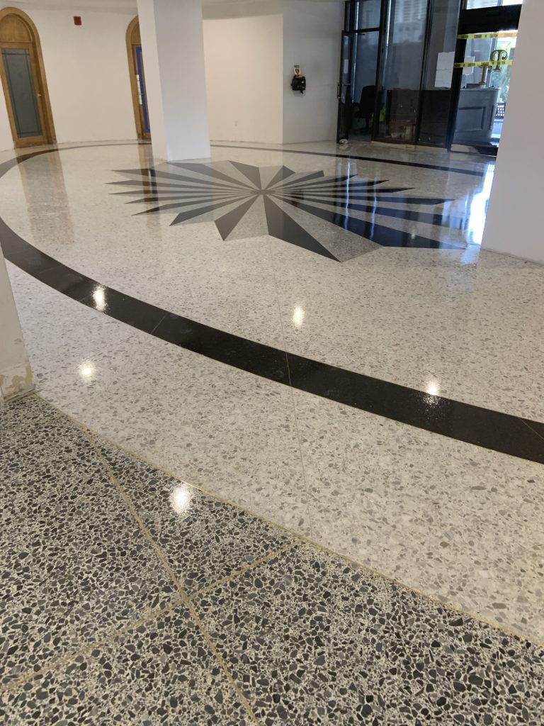 A polished epoxy flooring Palm Beach, FL features a large black geometric star center design, surrounded by curved black lines. Light terrazzo covers most of the area, with darker speckled flooring in the foreground.