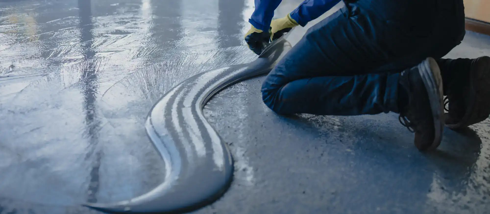 A person kneels on the ground in FL, wearing gloves, spreading wet concrete or epoxy flooring with a tool on a smooth surface in Miami-Dade or St. Lucie County, creating a wavy pattern.