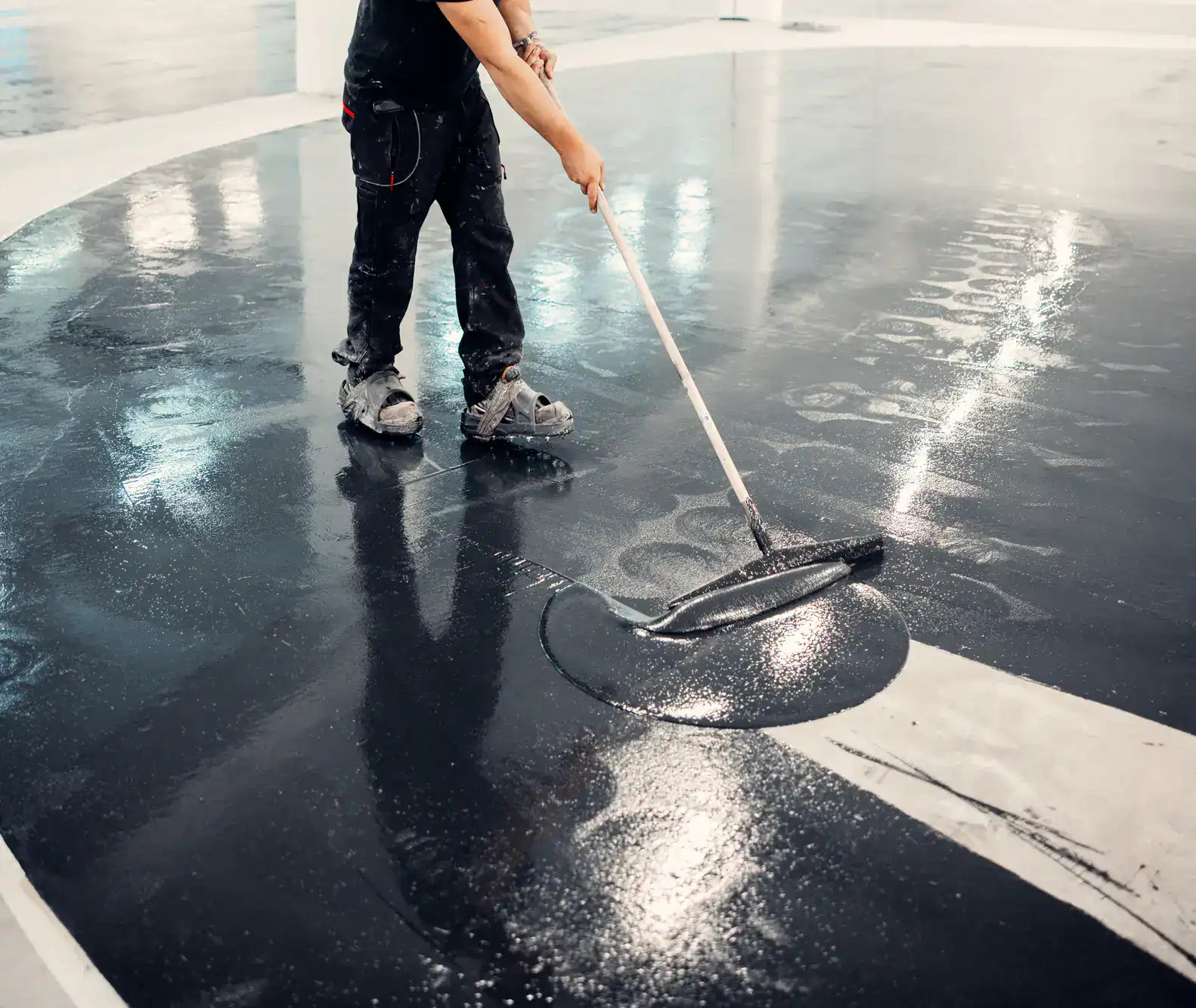 A person in St. Lucie County, FL uses a large roller to spread a shiny black coating over a concrete floor, working in a circular motion. The reflection of their legs and roller is visible on the wet surface.
