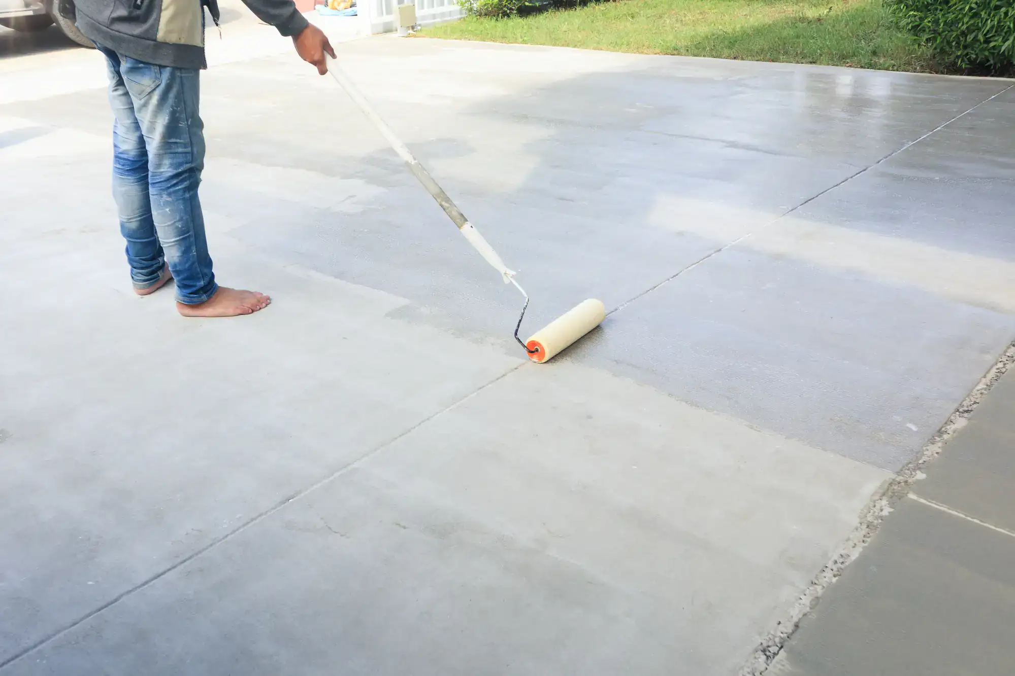 A person in jeans and a jacket applies a coat of paint or sealant to a concrete driveway using a long-handled roller, with grass visible in the background, perfect for homes in Broward and St. Lucie County, FL.