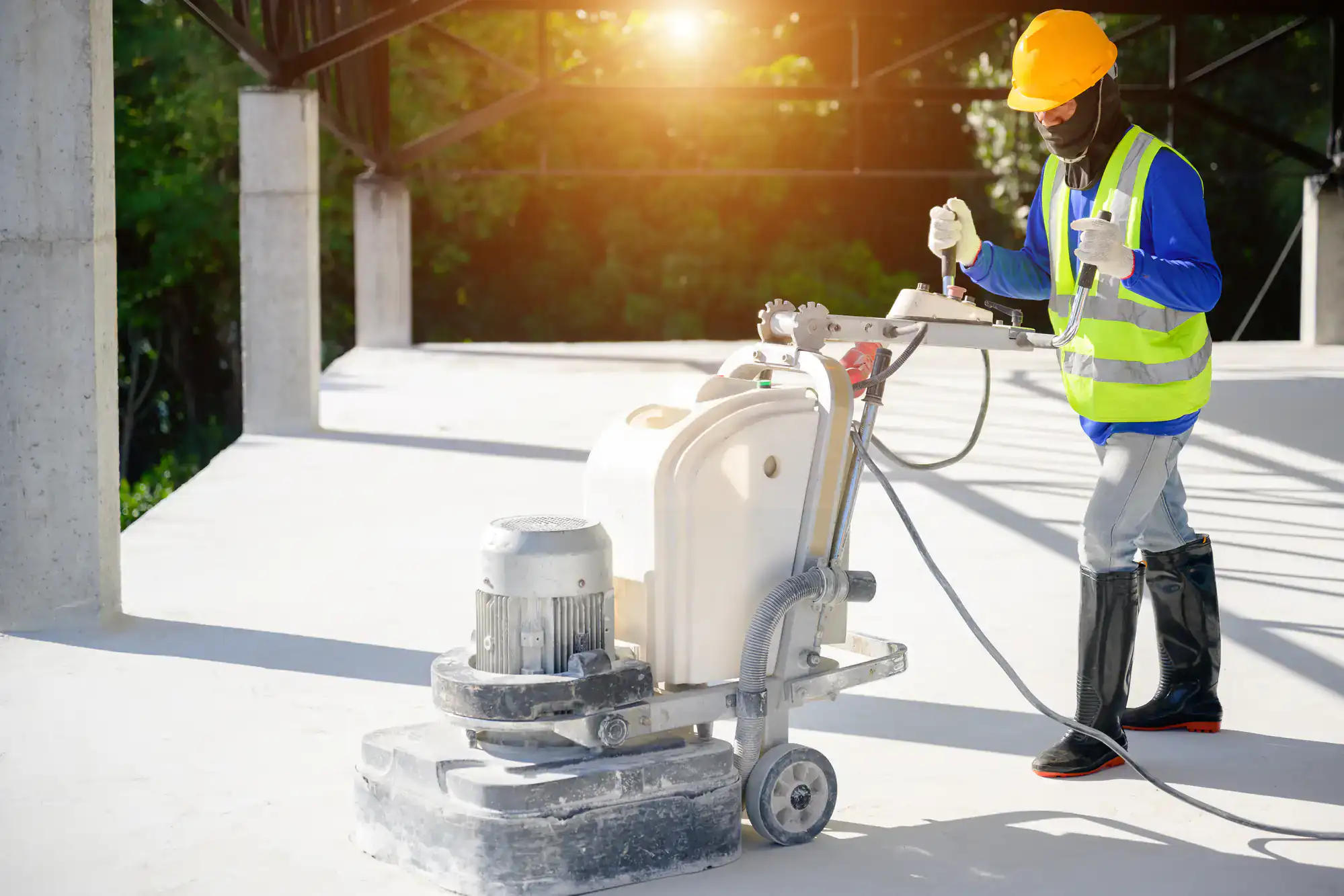 A construction worker in safety gear uses a floor polishing machine on a concrete surface inside an unfinished building in Miami-Dade, FL, with sunlight streaming through the background.