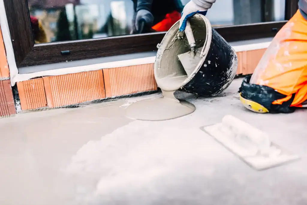 A construction worker in orange pants pours wet cement near a window, preparing a smooth surface for epoxy flooring Palm Beach. A trowel lies nearby on the ground.