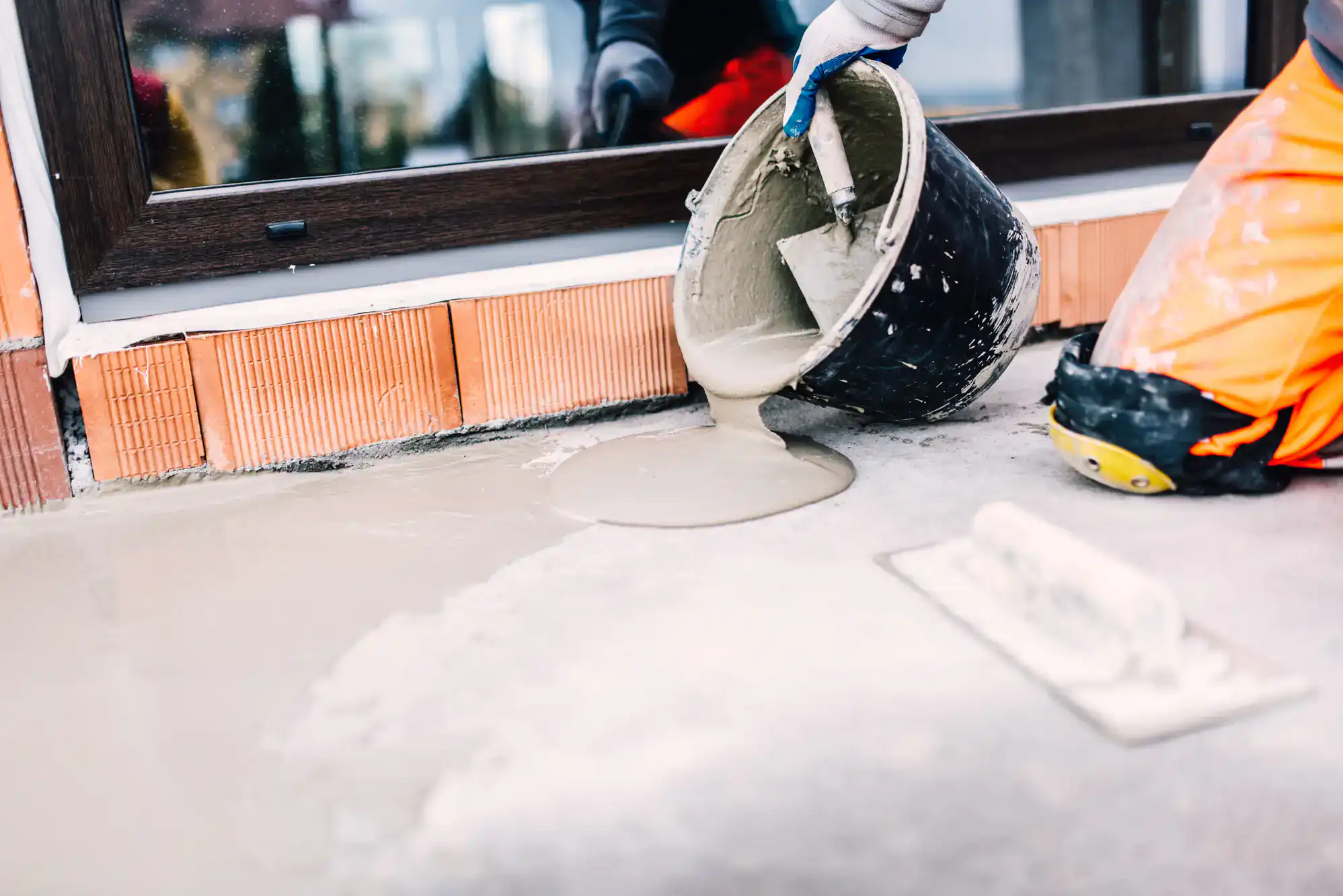 A construction worker in orange pants pours wet cement near a window, preparing a smooth surface for epoxy flooring Palm Beach. A trowel lies nearby on the ground.