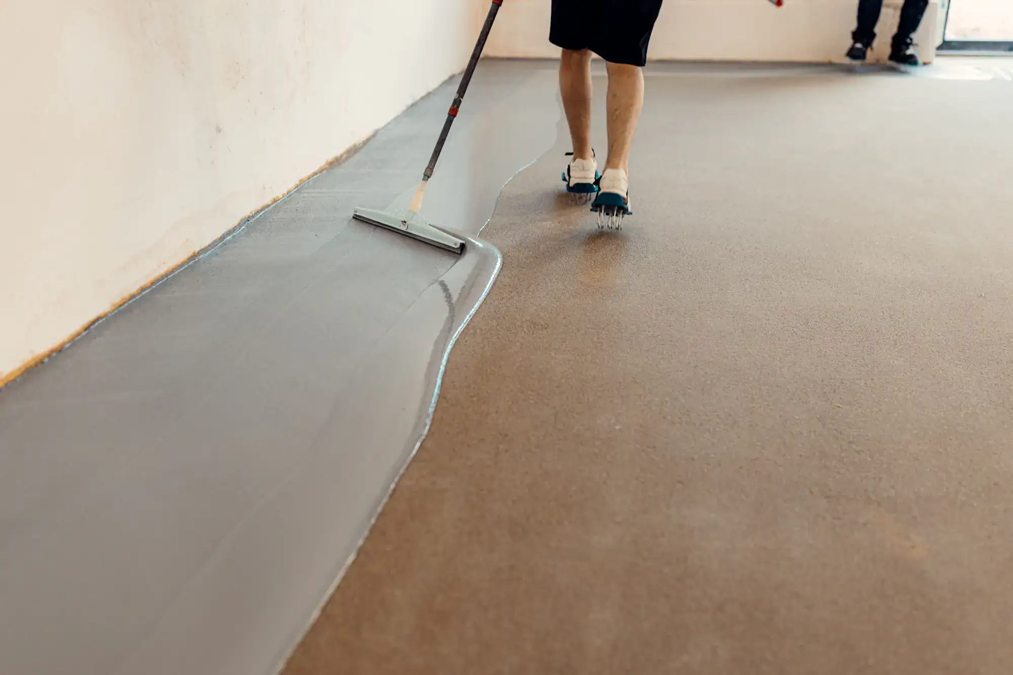 A person wearing spike shoes spreads a gray self-leveling floor compound, often used in epoxy flooring Palm Beach projects, with a long-handled tool over concrete. Another person stands in the background.