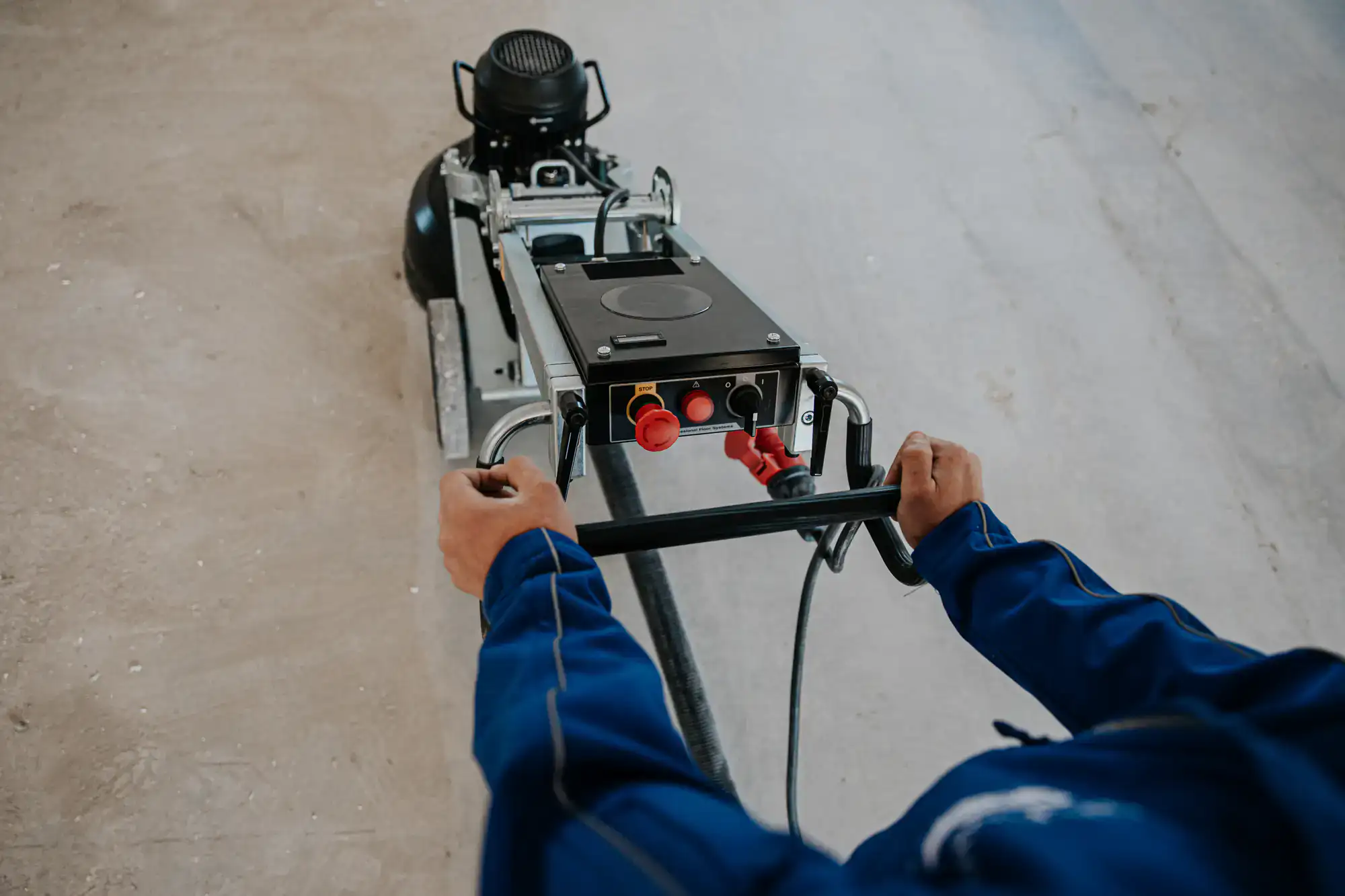 A person in blue workwear operates a large industrial floor cleaning machine on a bare concrete surface, servicing areas throughout Miami-Dade, Broward, and St. Lucie County.