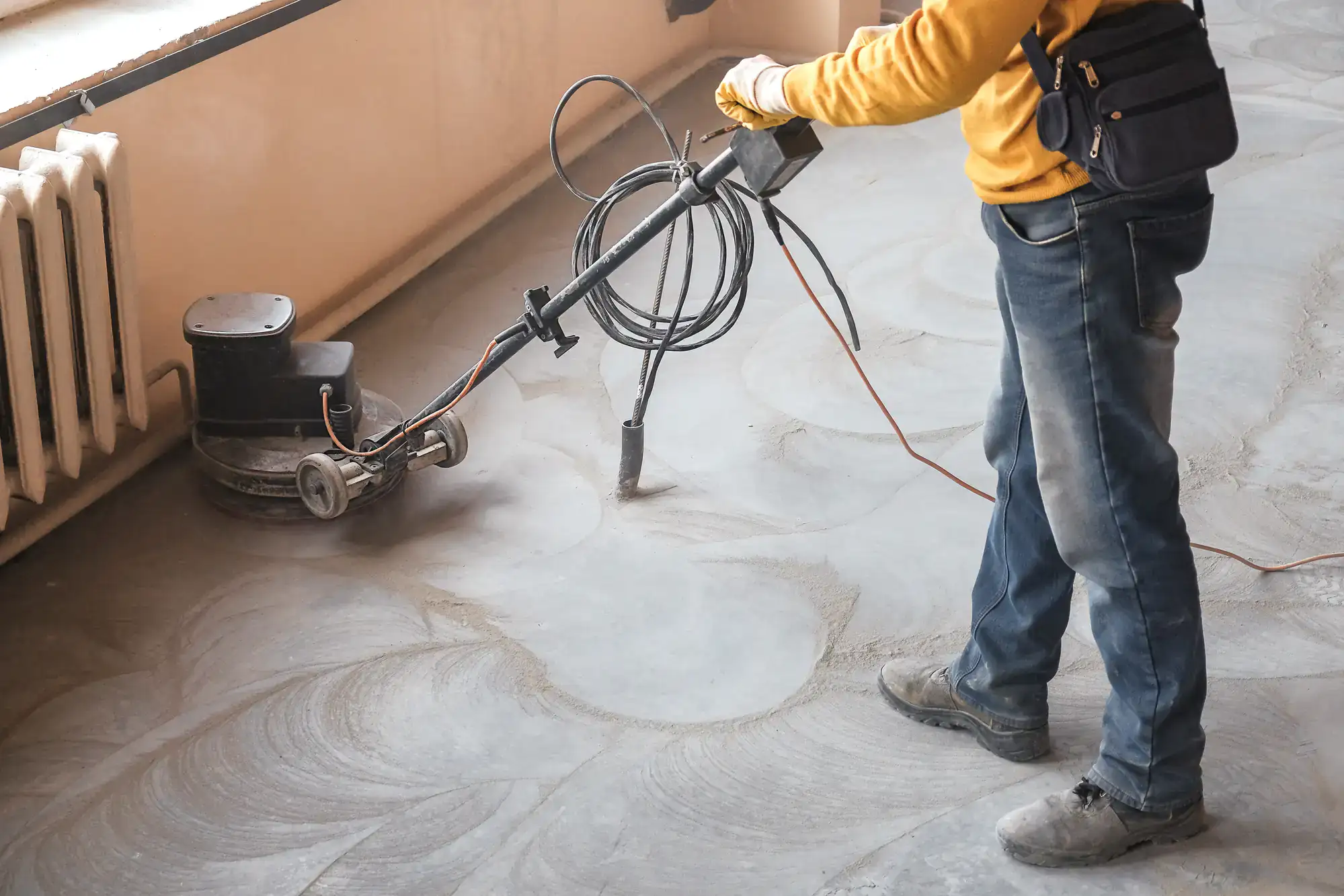 A person in jeans and a yellow shirt operates a floor polishing machine, smoothing a dusty concrete floor in an indoor room&mdash;a scene familiar to many epoxy flooring Palm Beach professionals&mdash;with a radiator visible on the left.