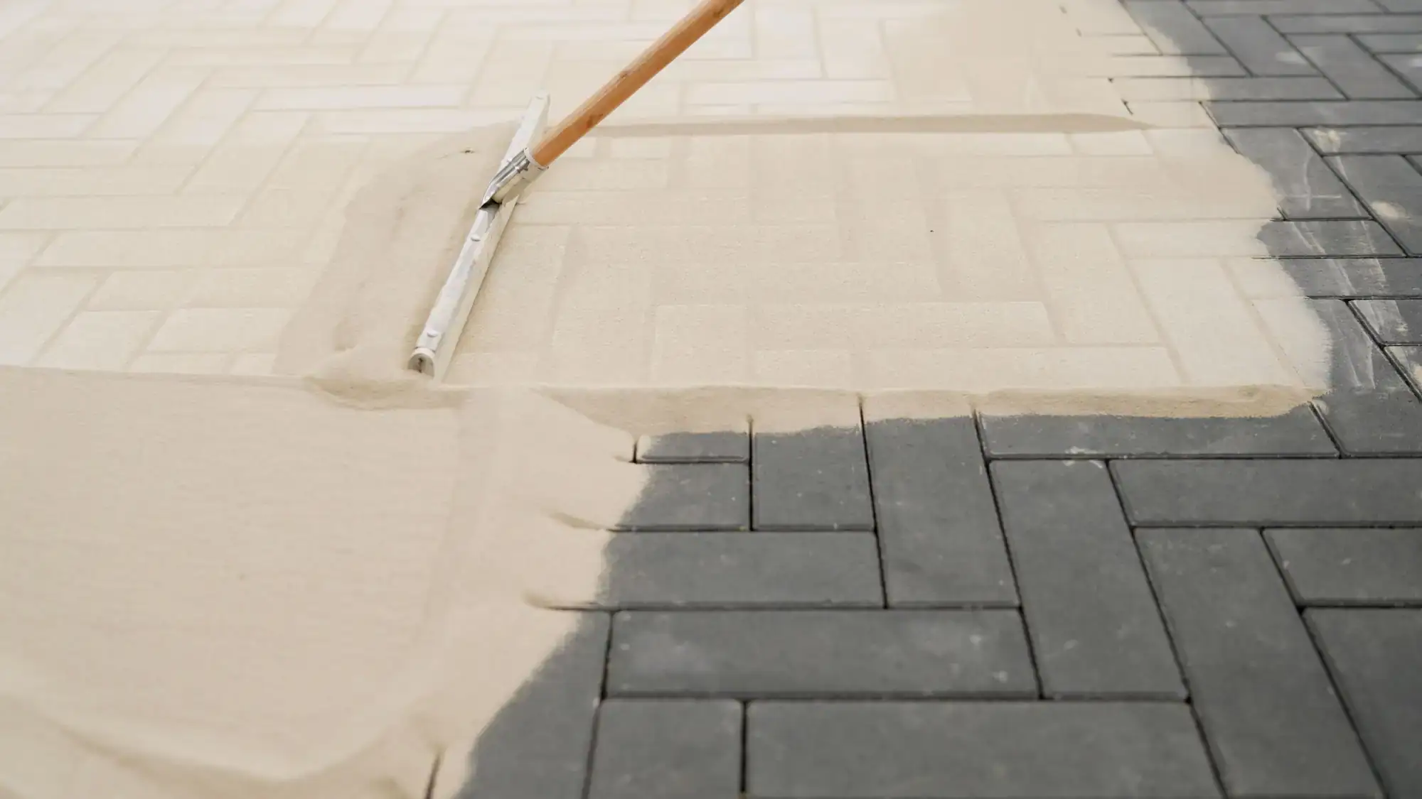 A rake spreads light-colored sand over dark gray brick pavers in a herringbone pattern, filling gaps for stability and a finished look, ideal for Miami-Dade or Broward hardscaping projects.