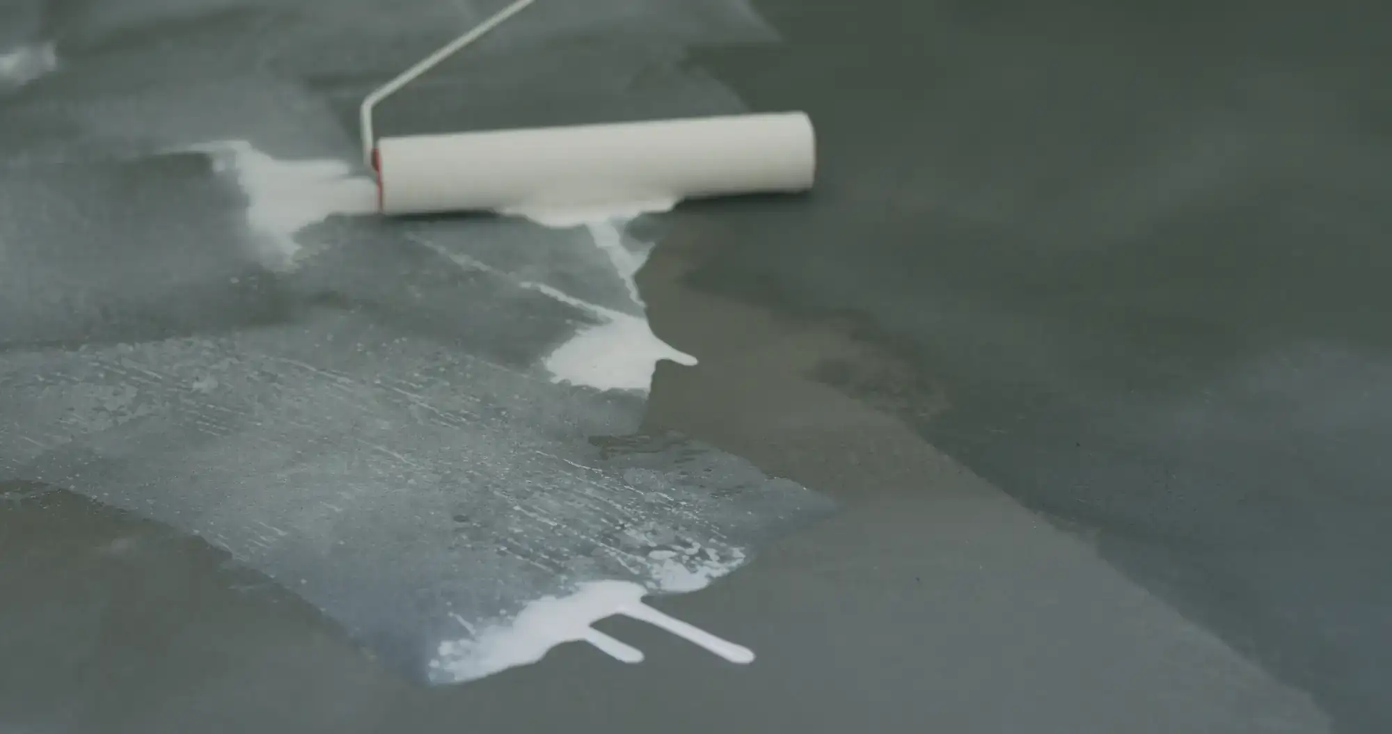 A paint roller applies a layer of white paint on a dark, smooth surface, with splashes and streaks of wet paint visible&mdash;perfect for refreshing epoxy flooring in Palm Beach.