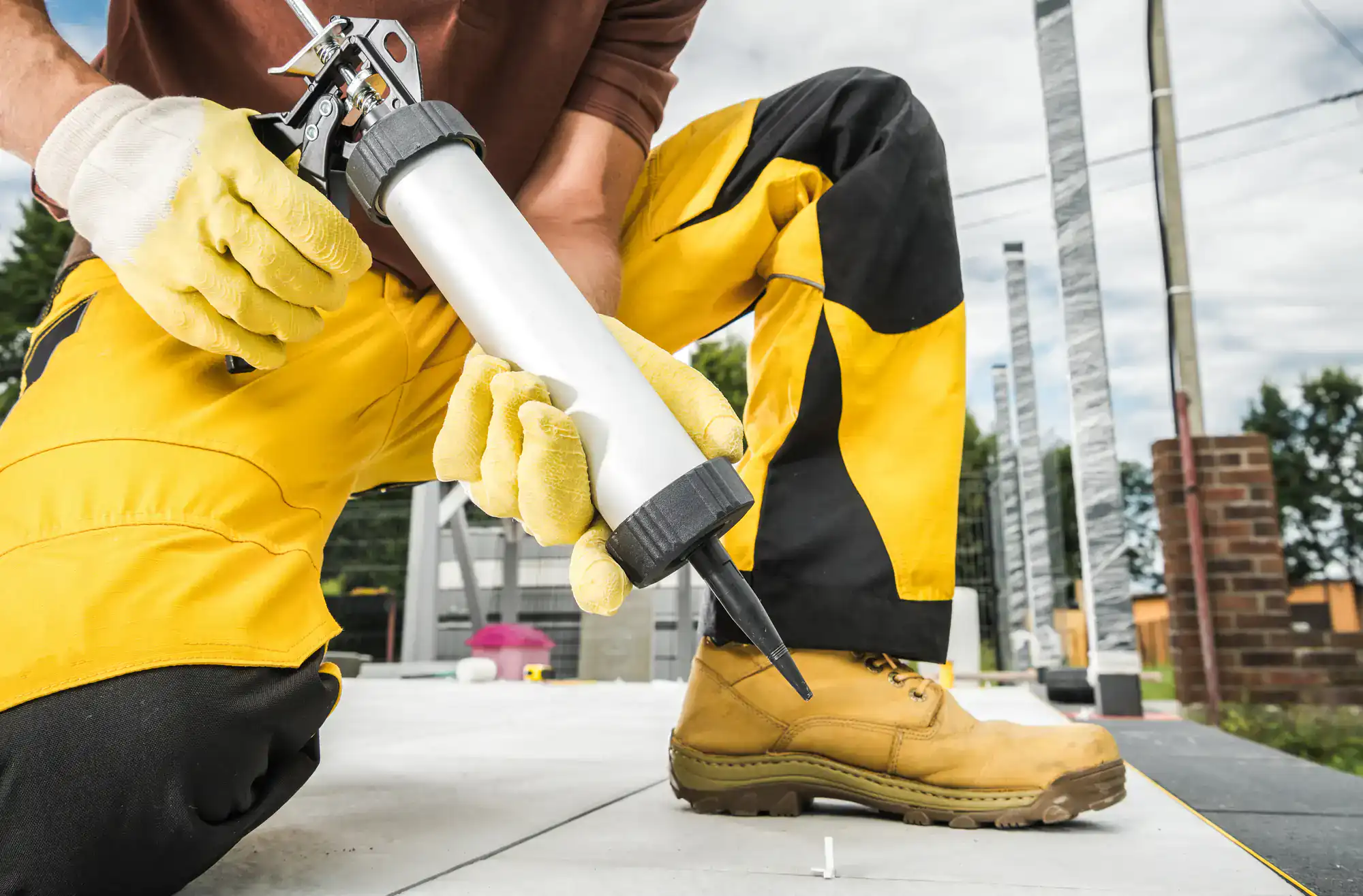 A worker wearing yellow gloves, yellow and black pants, and tan boots uses a caulking gun to apply sealant outdoors on a tiled surface—common prep work for epoxy flooring in areas like Palm Beach or Broward. Construction tools are visible in the background.