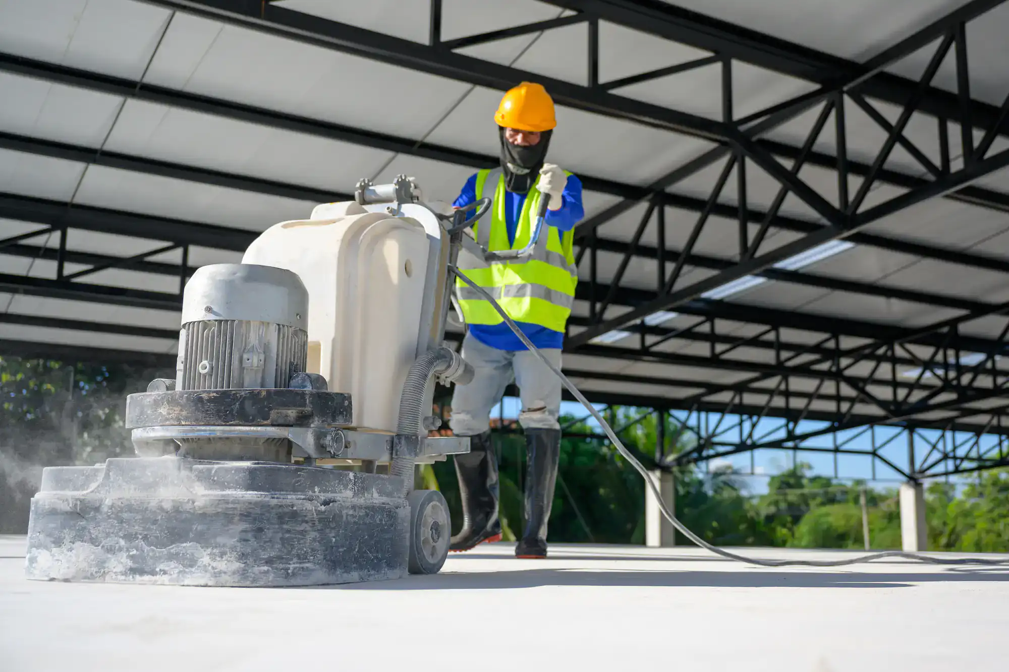A construction worker in a safety vest, helmet, and boots operates a floor grinding machine on a concrete surface, preparing for epoxy flooring Palm Beach inside a large, unfinished Miami-Dade, FL building with exposed steel beams.
