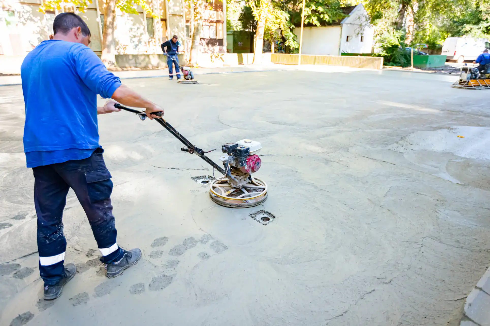 A worker in a blue shirt uses a power trowel to smooth wet concrete on a large outdoor surface in Broward. Other workers are visible in the background, with trees and buildings bordering the Miami-Dade and St. Lucie County site.