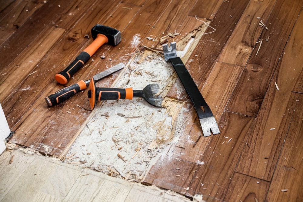 Three tools&mdash;a hammer, chisel, and pry bar&mdash;lie on a wooden floor in Broward, FL, with a section partially removed, exposing subfloor and wood debris scattered around.