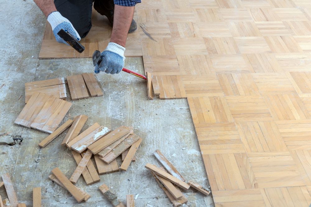 A person wearing gloves is installing or repairing parquet wooden flooring. Loose wooden tiles and a hammer are on the floor, with some tiles already removed and others being fitted into place.