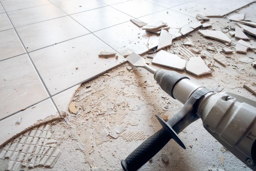 A power tool with a chisel attachment is being used to remove broken ceramic tiles from a floor in FL, leaving shattered pieces and dust scattered around the work area&mdash;readying the space for new epoxy flooring Palm Beach or Broward residents may desire.