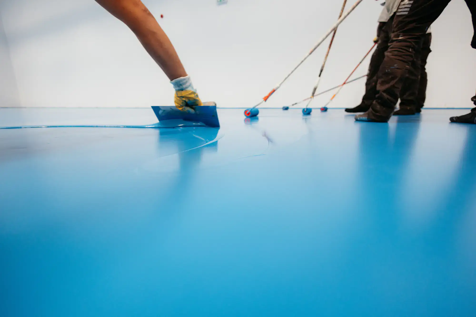Workers are applying a smooth blue epoxy coating to a floor using trowels and rollers. The image focuses on gloved hands spreading the coating, with additional workers visible in the background.