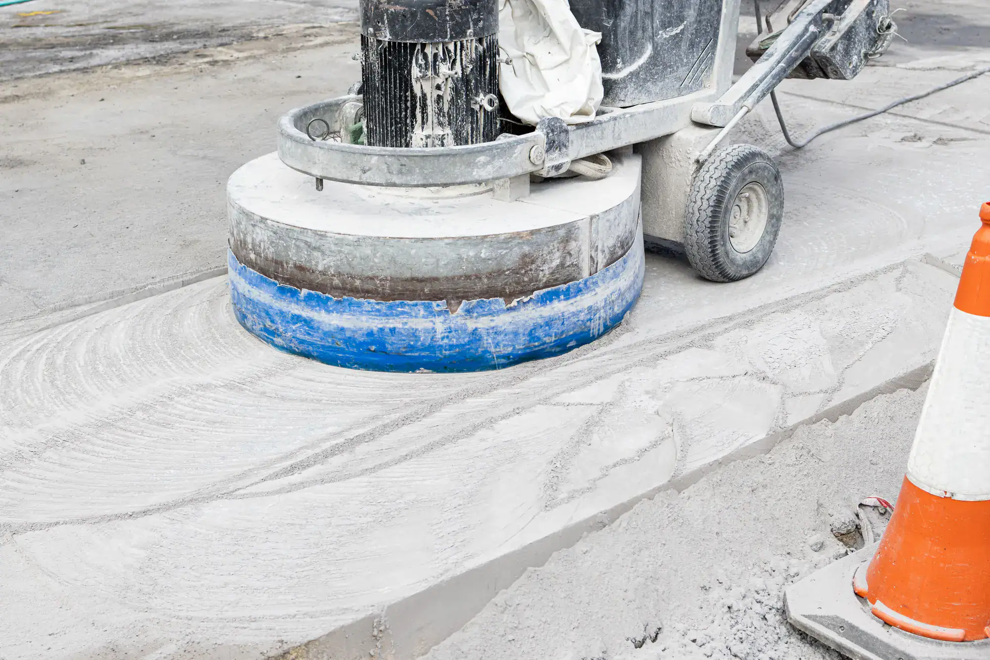 A close-up of a concrete polishing machine smoothing a concrete floor, with white dust and a traffic cone visible nearby.