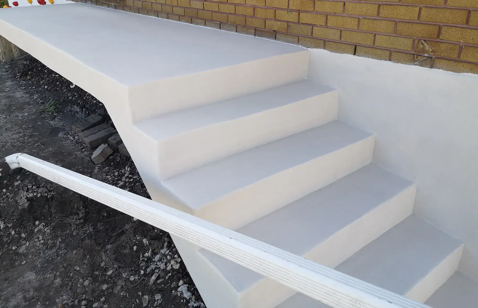 Concrete steps with smooth, white finish leading up to a raised platform beside a brick wall, with gravel and soil on the surrounding ground. A white handrail runs along one side.