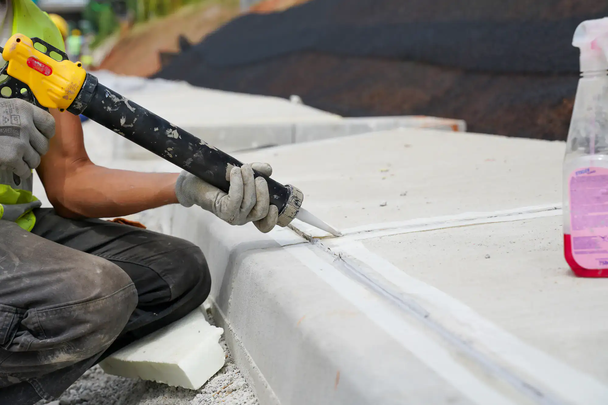 A worker applies sealant to the gap between concrete sections using a caulking gun. The person wears gloves, and construction materials are visible around the site.