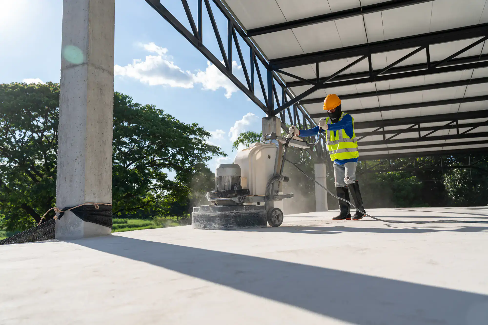 A construction worker wearing a helmet, safety vest, and boots operates a floor polishing machine on a concrete surface inside an open steel structure, with trees and blue sky visible in the background.