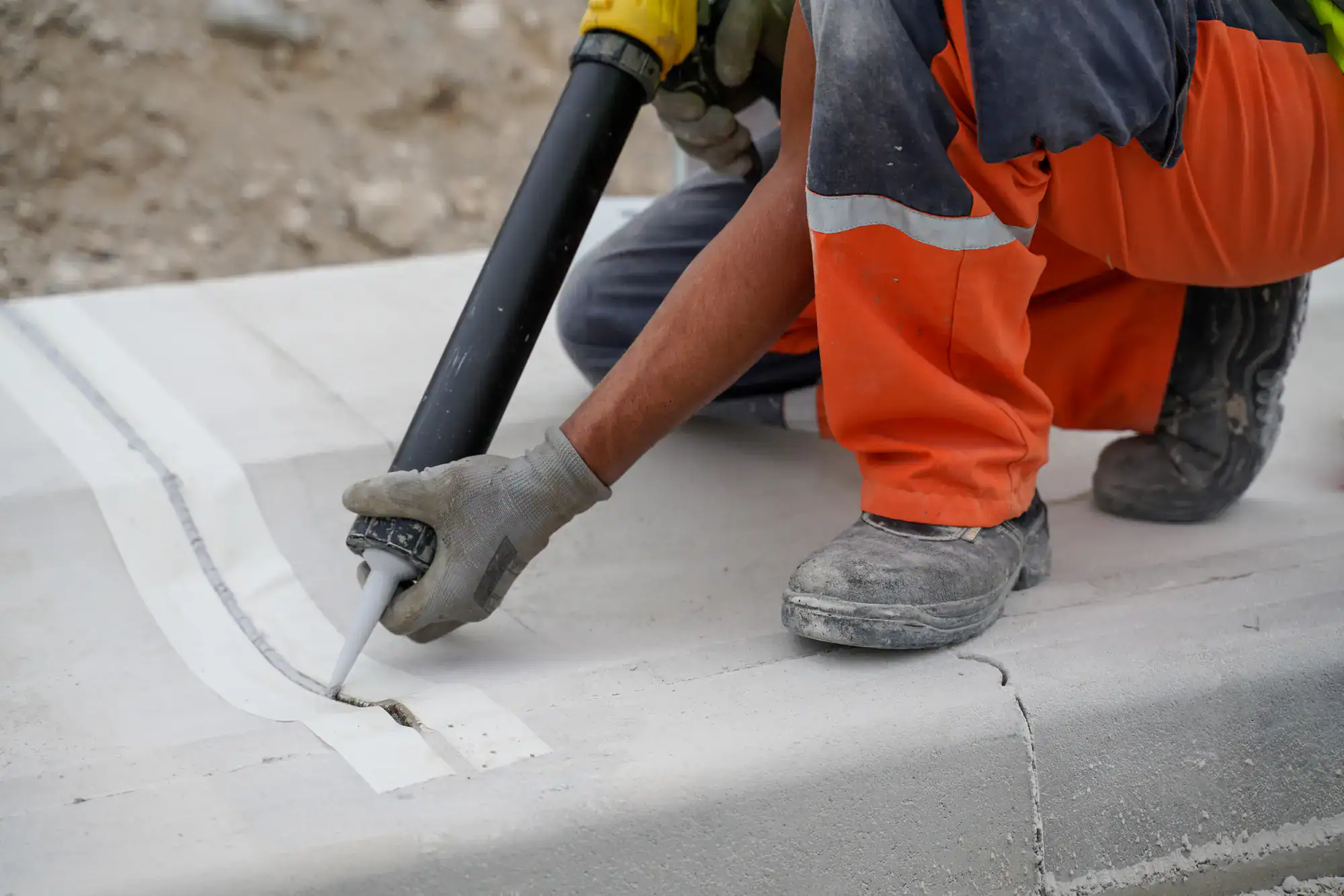 A construction worker wearing orange pants and gloves applies sealant to a crack in a concrete surface using a caulking gun. The area appears to be part of a road or sidewalk under repair.