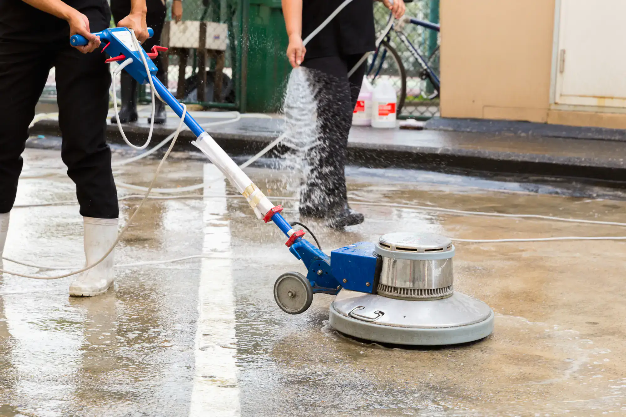A person using a blue and silver floor cleaning machine on a wet outdoor surface, while another person sprays water nearby; both wear black clothes and rubber boots.