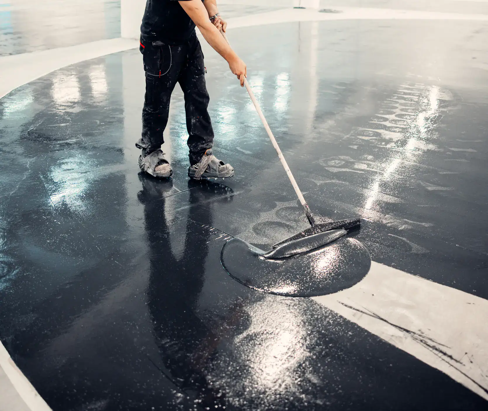 A person wearing work clothes uses a large squeegee to spread a dark, shiny coating on a concrete floor inside a building, possibly applying epoxy or sealant. Only the person&rsquo;s legs and arms are visible.