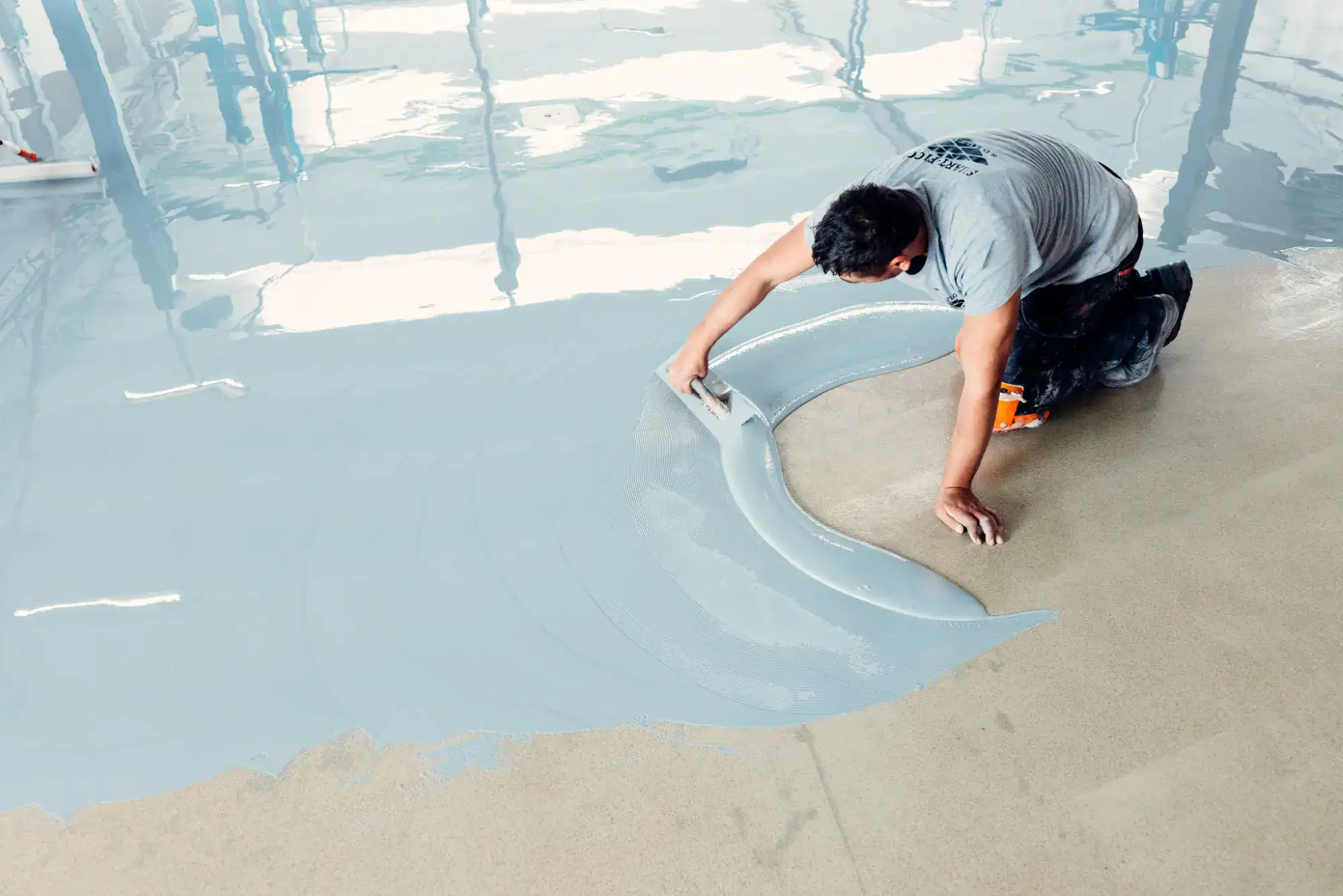 A person kneeling on the floor uses a trowel to spread a smooth, gray epoxy coating over a concrete surface in a large, bright room.
