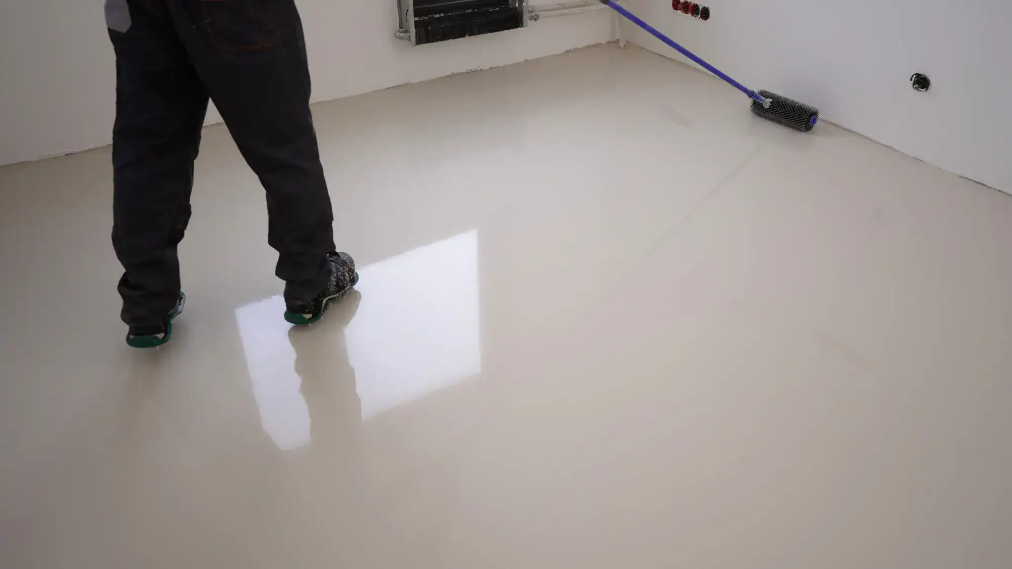 A person stands on a shiny, freshly laid floor, likely applying or inspecting a smooth epoxy or resin coating in a bright, empty room with white walls and some visible tools.