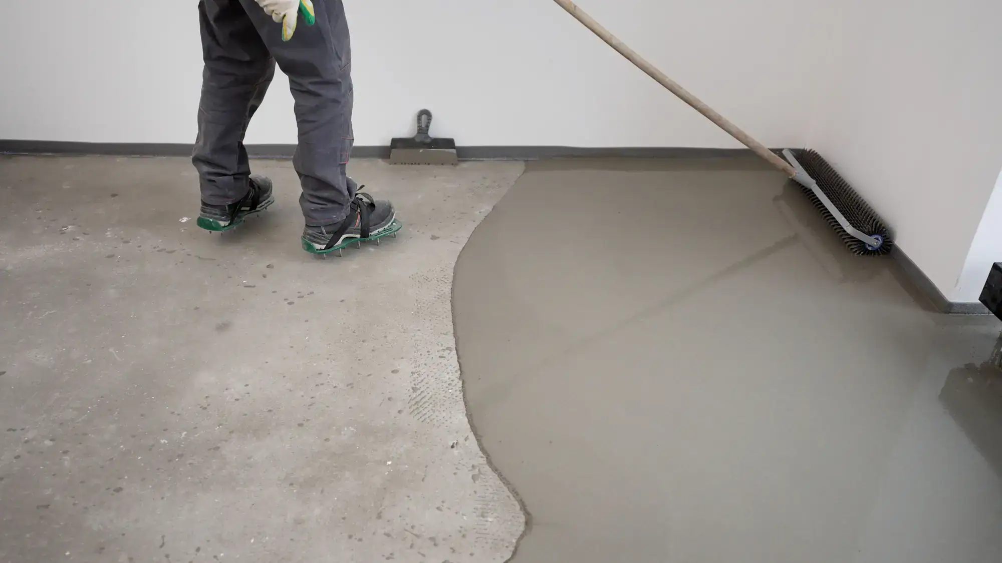 A person in work pants and green shoes uses a broom to spread wet concrete or floor leveling compound over a bare concrete floor near a white wall.