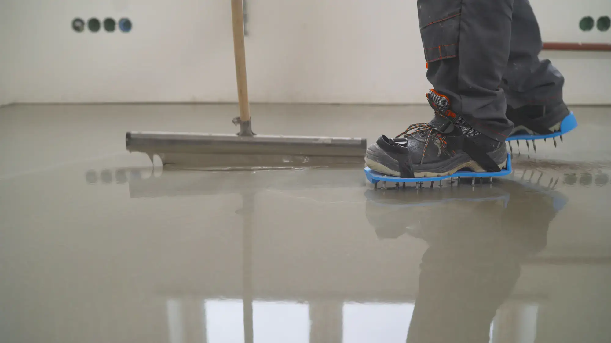 A worker wearing spiked shoes uses a tool to spread and level wet concrete or floor screed in an unfinished room.