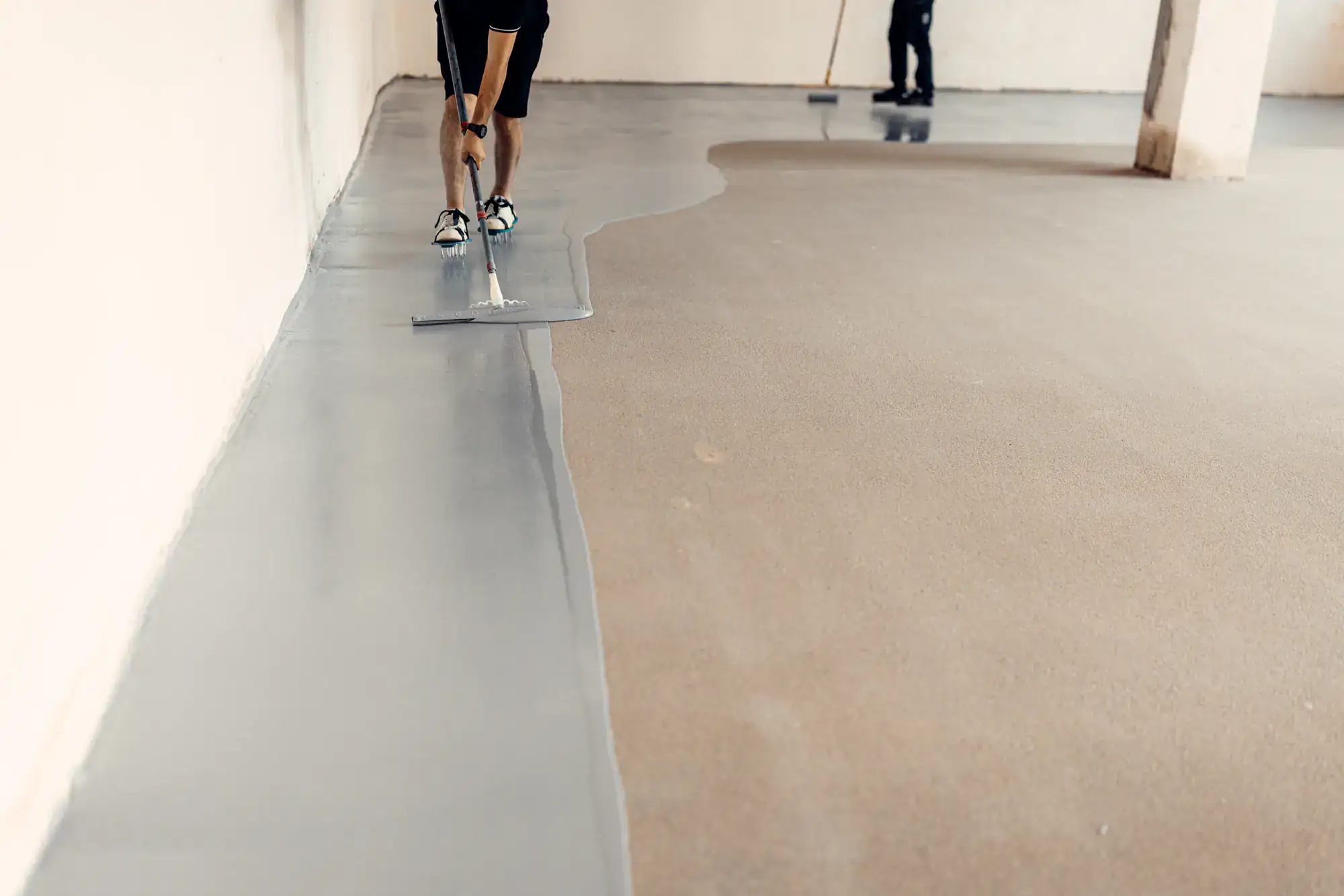 Two people apply a smooth, grey epoxy coating to a concrete floor in an indoor space, using large floor squeegees. One person works in the foreground, while another works further back near a column.
