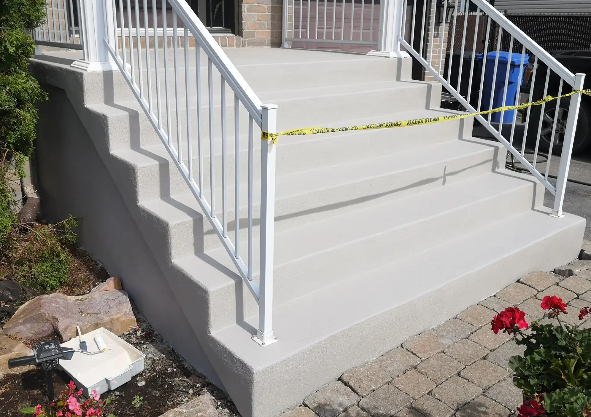 Freshly painted concrete stairs with white railings, blocked off by yellow caution tape. Painting supplies and a white tray are on the ground nearby, next to pavers and red flowers.