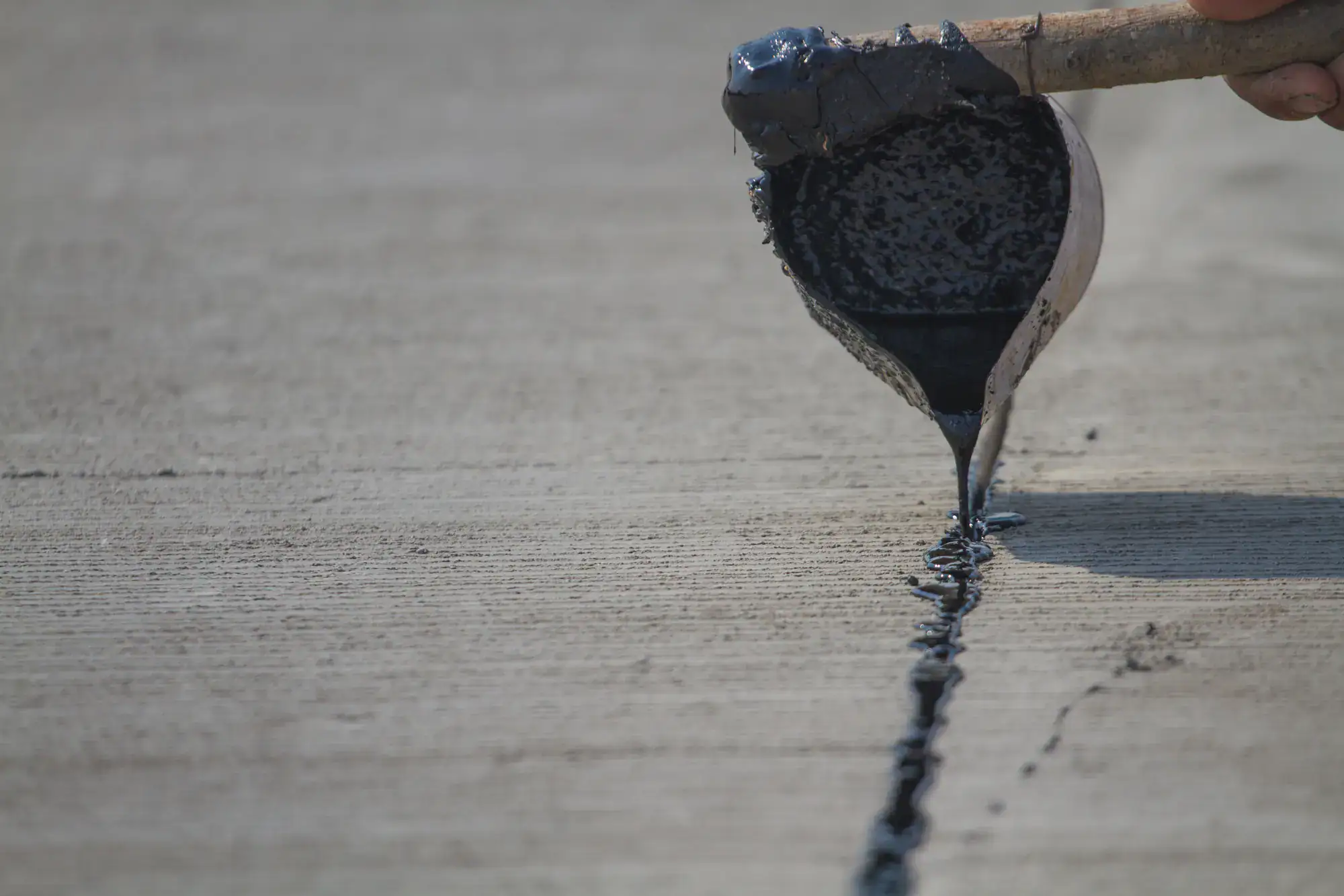 A close-up of a hand pouring black liquid sealant from a ladle into a crack on a concrete surface for repair.