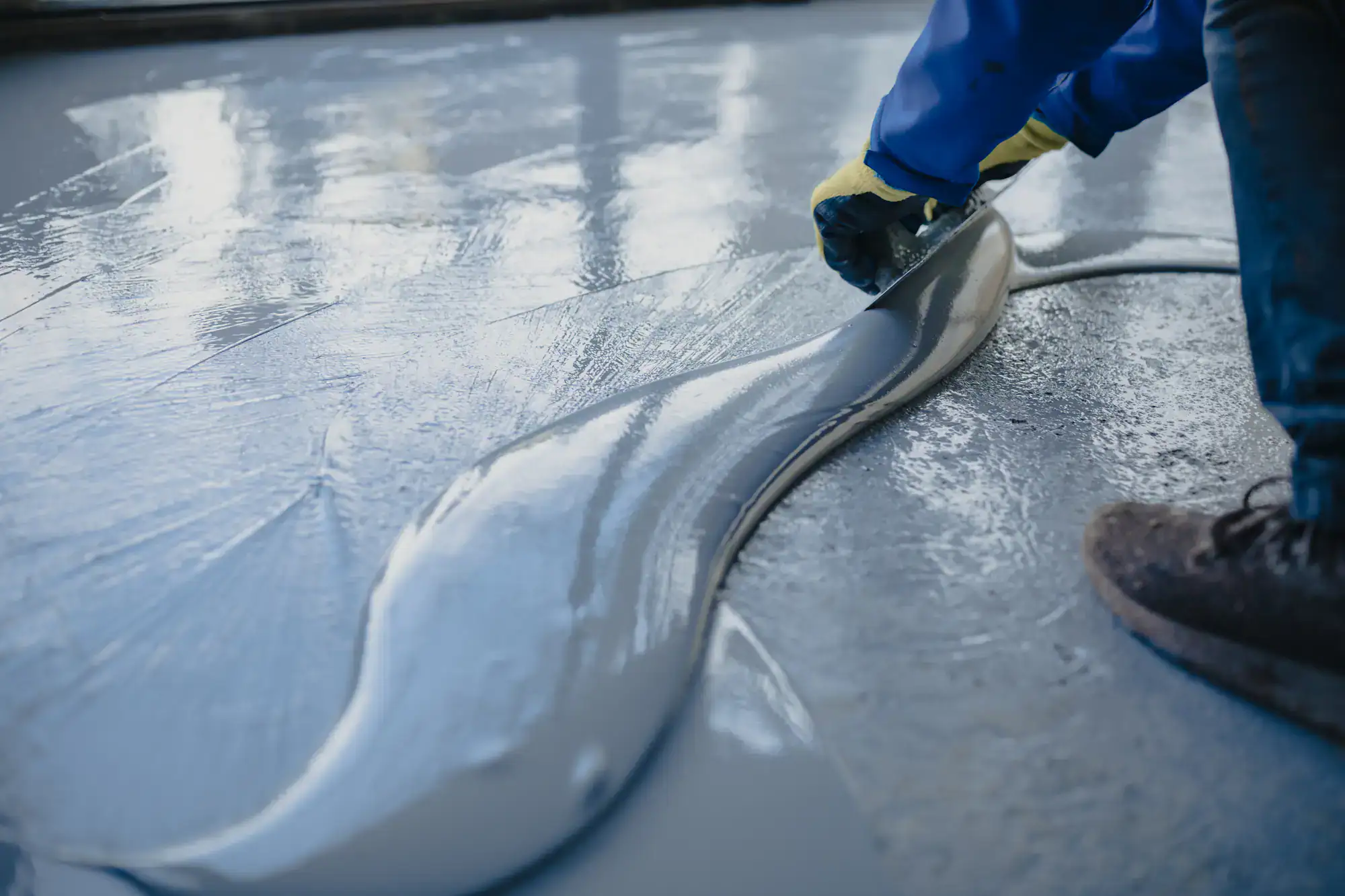 A person wearing gloves and boots uses a trowel to spread wet, gray self-leveling compound over a concrete floor for smoothing and resurfacing.