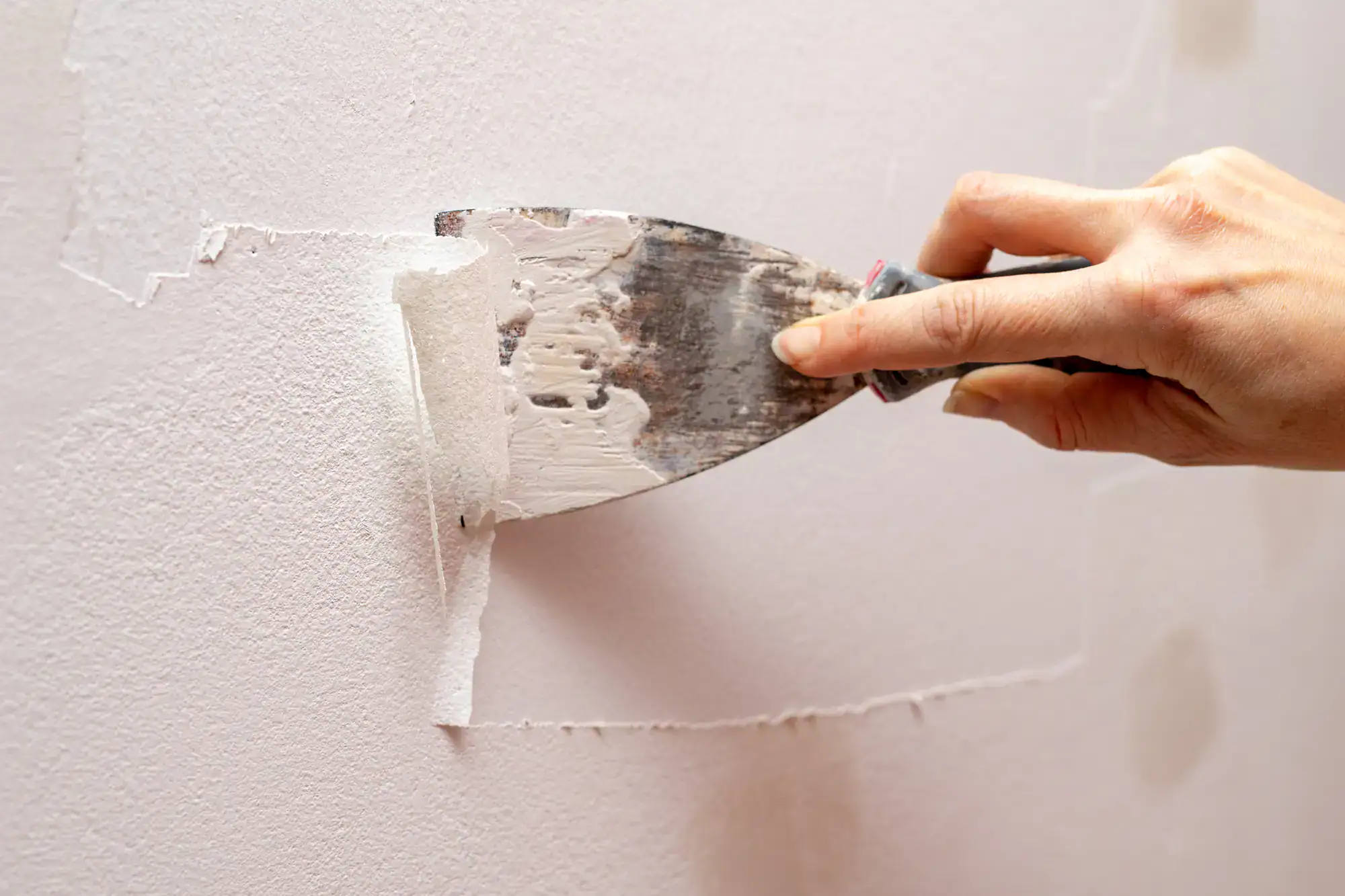 A hand uses a metal putty knife to spread white spackle on a pale pink wall, smoothing out imperfections in the surface.