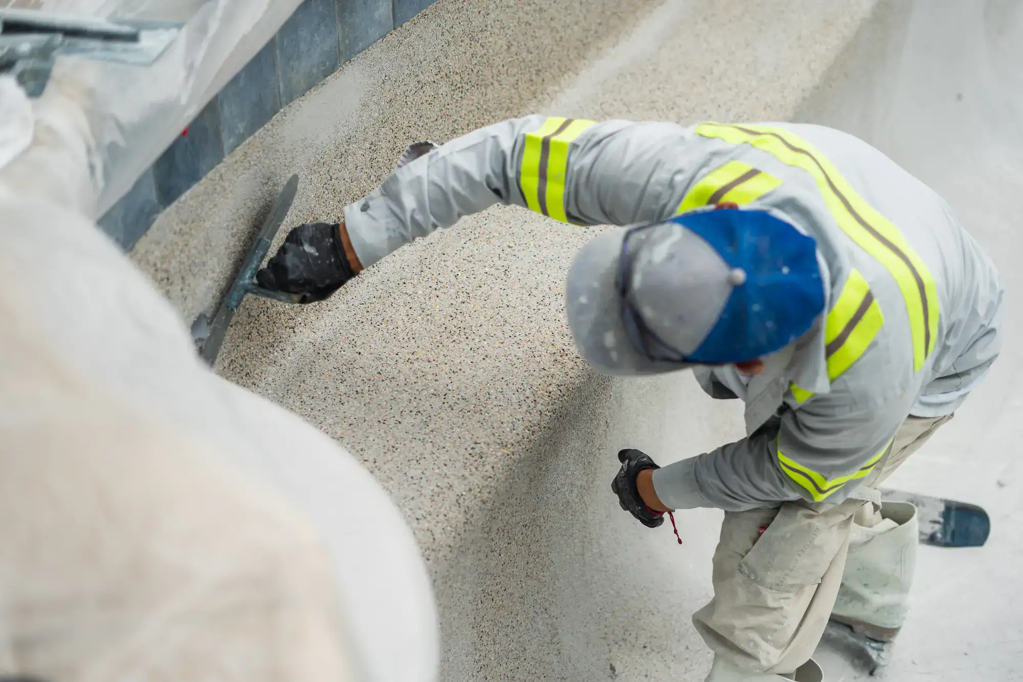 A worker in a gray jacket with yellow reflective stripes smooths the surface of a concrete pool wall with a trowel, wearing a blue and gray cap and black gloves.