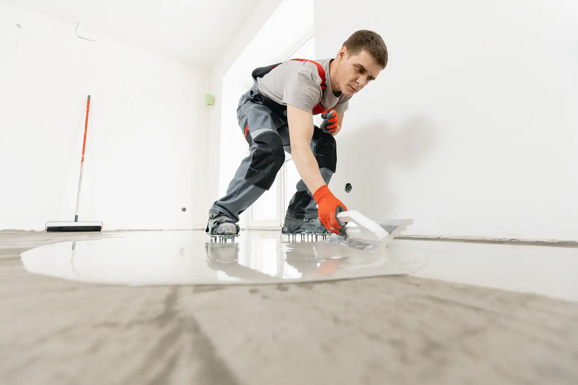 A construction worker wearing gloves and protective gear smooths wet concrete on a floor in a bright, unfinished room using a trowel.