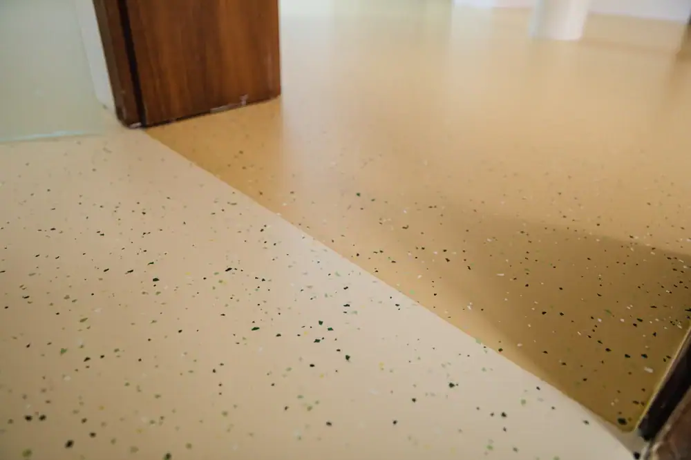 Close-up view of a smooth, beige, speckled epoxy floor surface near a wooden door frame and white wall in an indoor space. The floor features small flecks in black, green, and white.