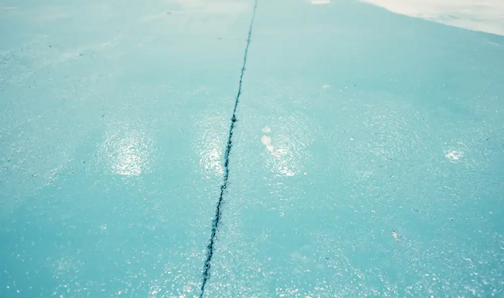 A close-up view of a light blue concrete surface with a single visible crack running vertically through the center. The surface appears glossy and slightly wet, reflecting nearby light.