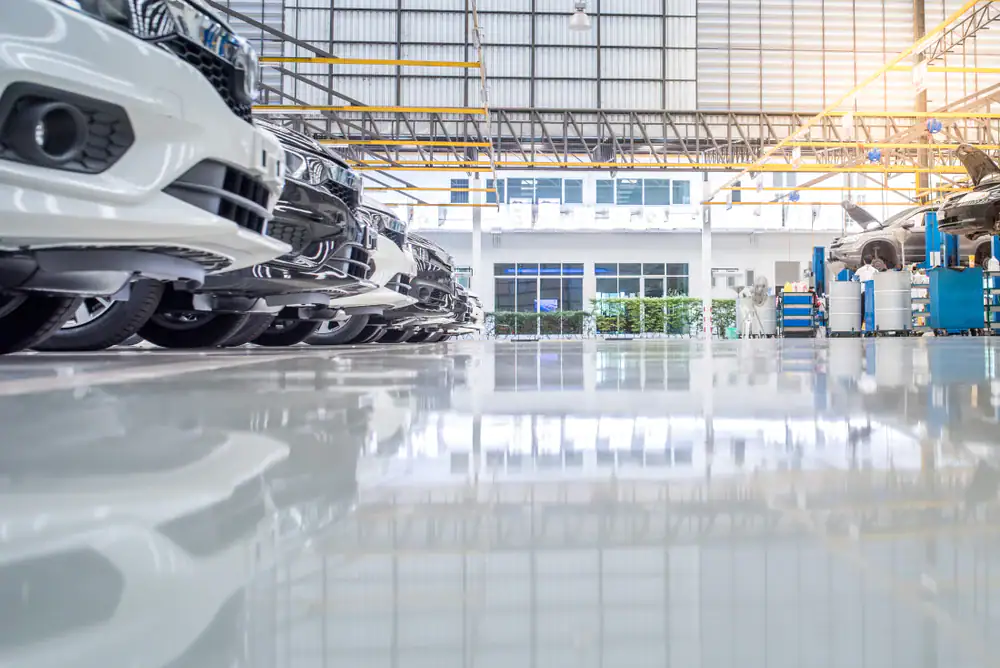A row of new cars is parked inside a clean, bright, spacious automotive factory or service center, with large windows, high ceilings, and industrial equipment visible in the background.